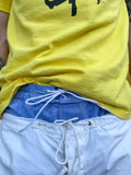 Close-up of light blue chambray boxer shorts with a board shorts-inspired elastic waistband and white drawstring, layered above white trouser and beneath a yellow T-shirt.