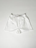 White cotton boxer shorts with a board shorts-inspired design, featuring an elastic waistband, white drawstring, and relaxed fit.