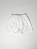 White cotton boxer shorts with a board shorts-inspired design, featuring an elastic waistband, white drawstring, and relaxed fit.
