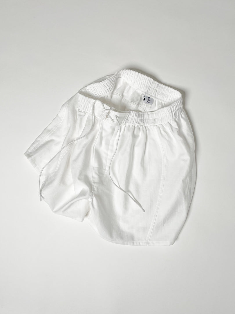 White cotton boxer shorts with a board shorts-inspired design, featuring an elastic waistband, white drawstring, and relaxed fit.