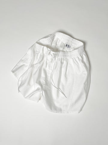 White cotton boxer shorts with a board shorts-inspired design, featuring an elastic waistband, white drawstring, and relaxed fit.